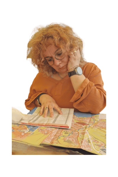Eine Frau mit orangefarbenen Oberteil sitzt nachdenklich vor einer Landkarte und einem Fahrplan. Es symbolisiert wiederholte Long Covid-Rückfälle und gewonnene Erfahrung, weil sie weiß, dass es einen Weg raus gibt.