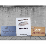 Drei Protestplakate für die Liegenddemo ME/CFS lehnen an einer rissigen grauen Betonwand mit davor angrenzenden Pflastersteinen. Die Forderung auf den Plakaten lautet: Anerkennung, Versorgung und Forschung zum ME/CFS und LongCovid