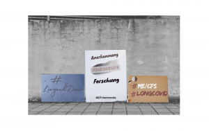 Drei Protestplakate für die Liegenddemo ME/CFS lehnen an einer rissigen grauen Betonwand mit davor angrenzenden Pflastersteinen. Die Forderung auf den Plakaten lautet: Anerkennung, Versorgung und Forschung zum ME/CFS und LongCovid